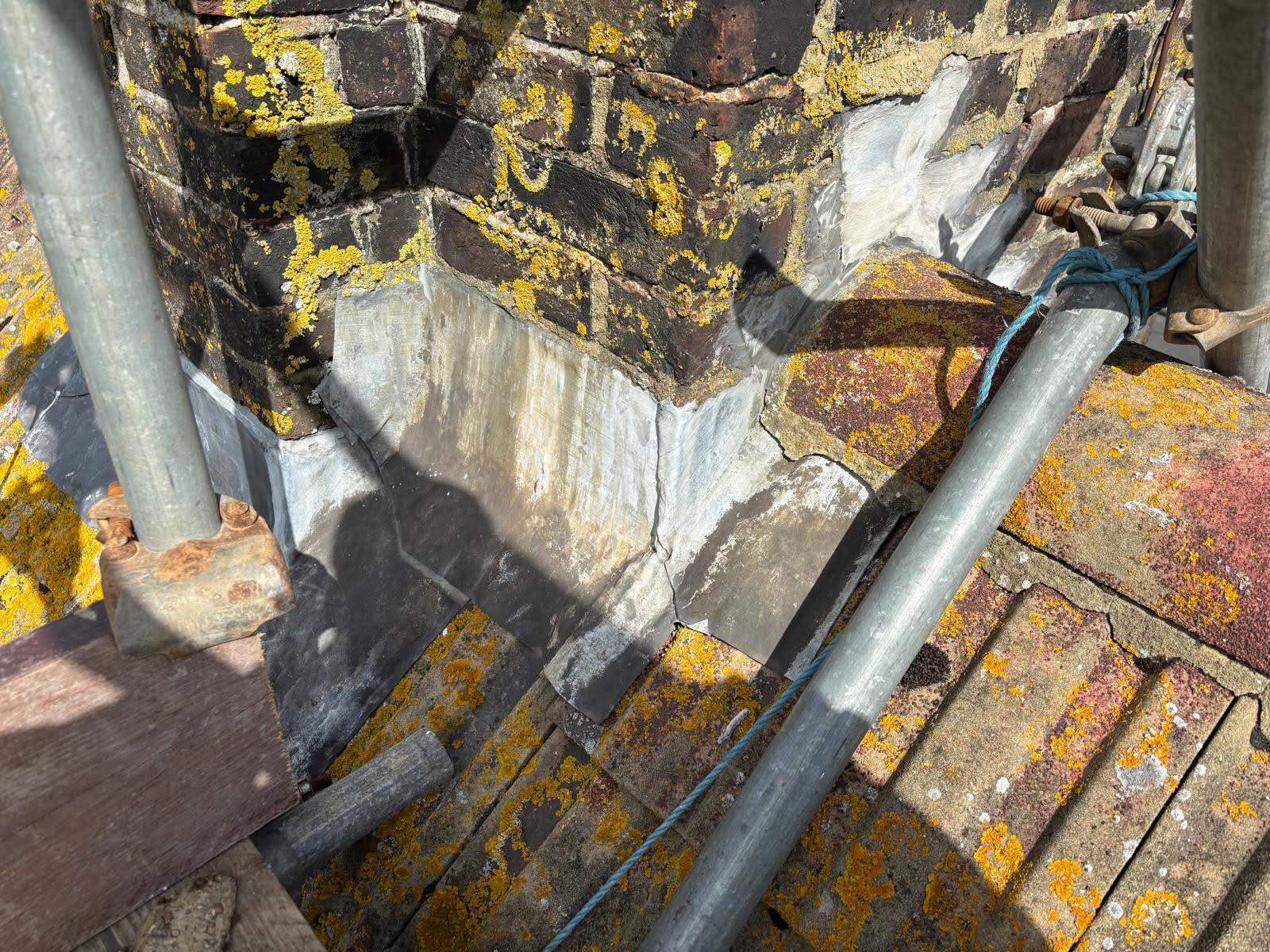 Close-up of damaged old lead flashing at chimney base — cracked and lifted sections allowing water ingress behind the brickwork